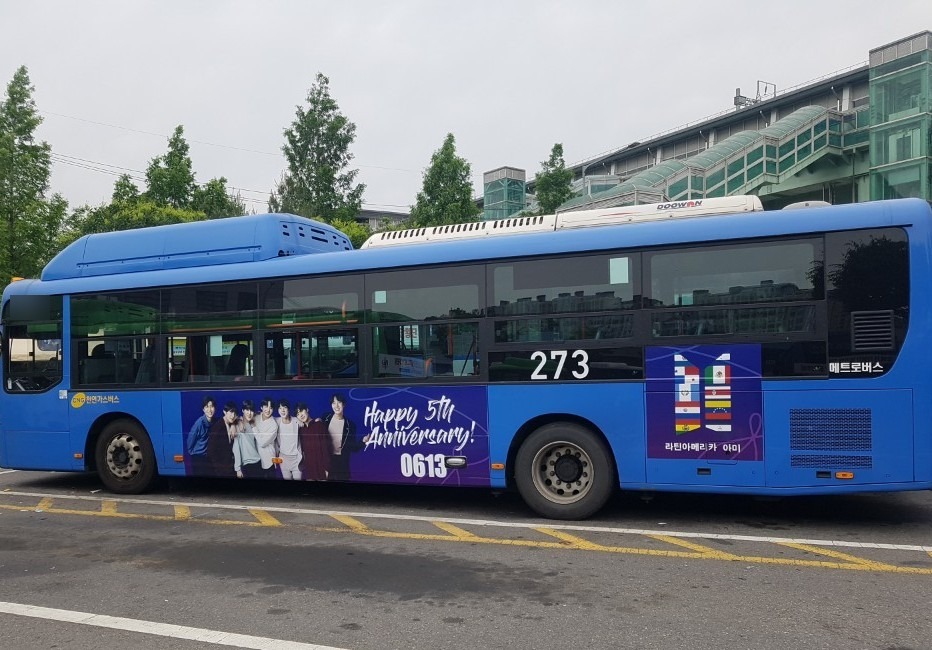 BTS anniversary ad on the road side of a blue Seoul city bus, a purple panel with a group photo and the words "Happy 5th Anniversary! 0613"