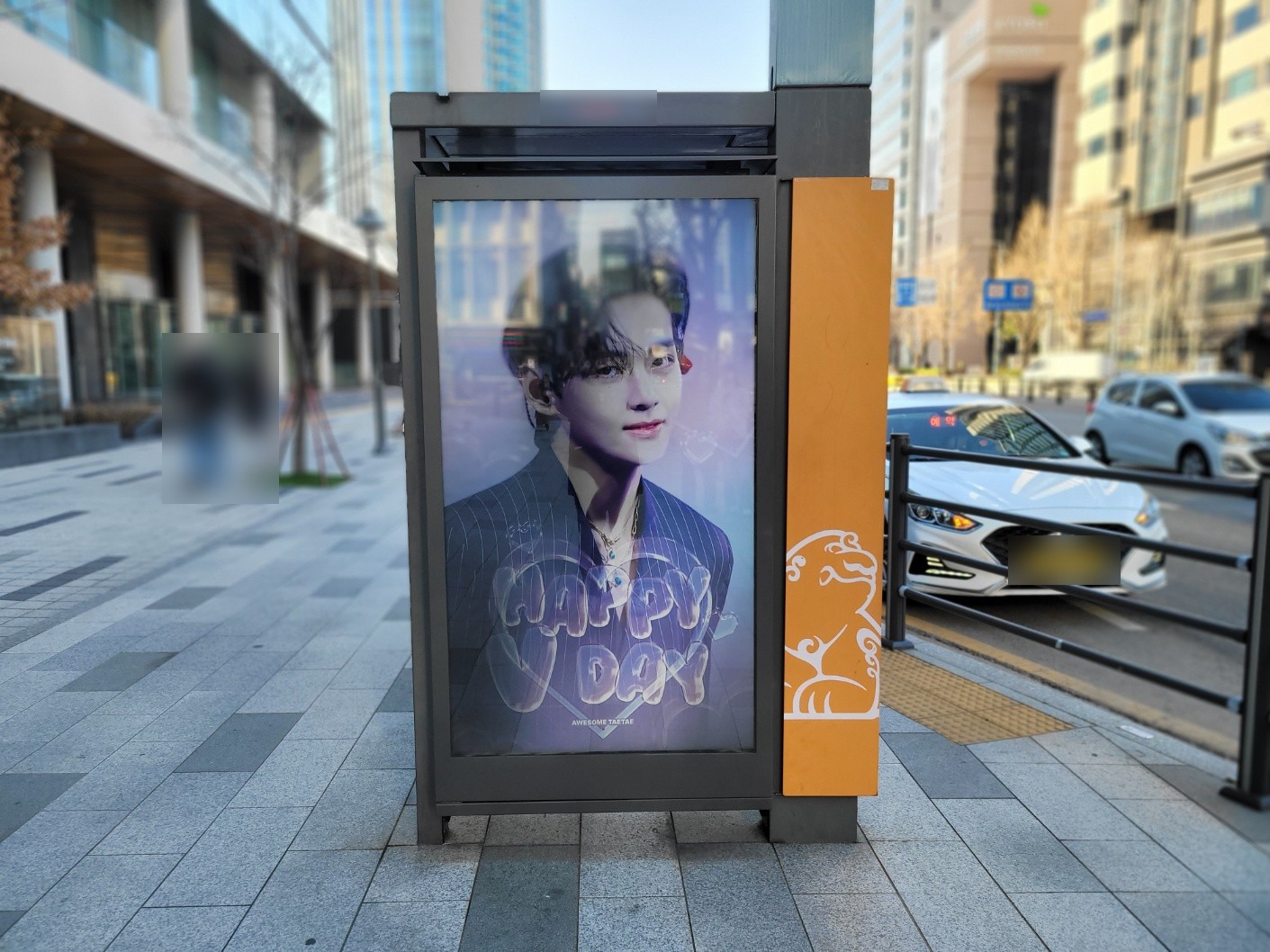 BTS V birthday ad on an upright digital screen at a street shelter in Seoul, showing a portrait in a pinstripe suit and the words HAPPY V DAY