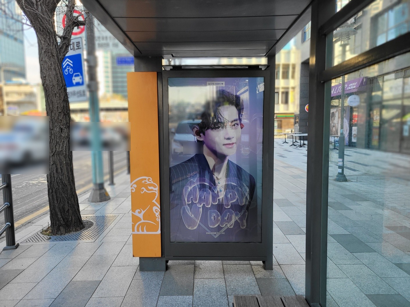 Screen at a shelter beside a street tree and road signs, showing the violet suit design with the credit line under the heart