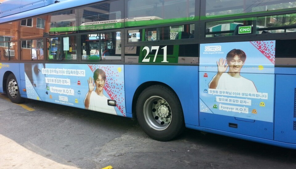 Jang Woo-hyuk birthday ad on the side of a blue Seoul city bus, sky-blue panels with a waving photo, Korean birthday text and small pixel-game characters