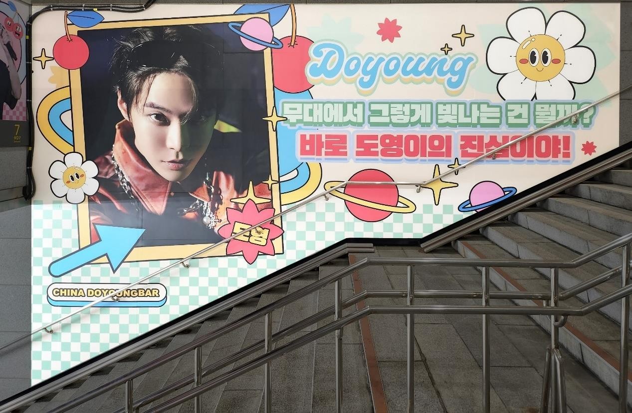 Stairway wall with a framed photo on the left, "Doyoung" in blue script, a two-line Korean message and cartoon planets and flowers on a checked background