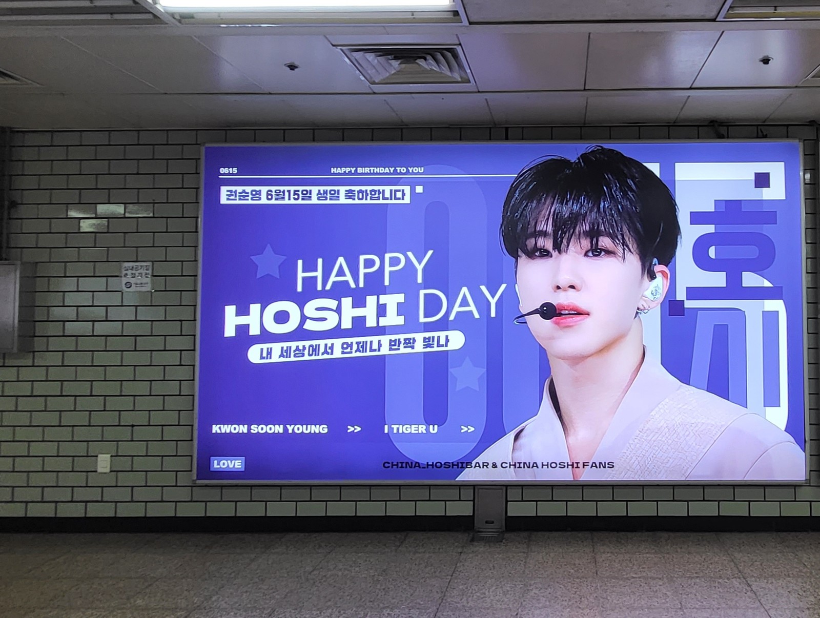 The whole lightbox on a white brick wall in a station passage, with the fan credit along the bottom edge